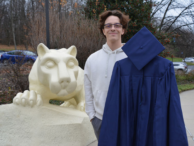 A male student holding academic regalia and standing next to the Nittany Lion Shrine at Penn State York