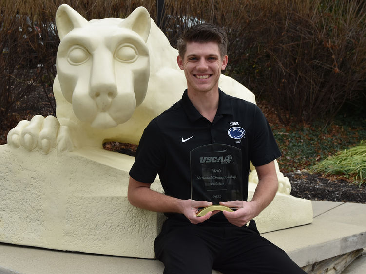 Male student holding golf award as he sits  in front of the Penn State NIttany Lion Shrine 