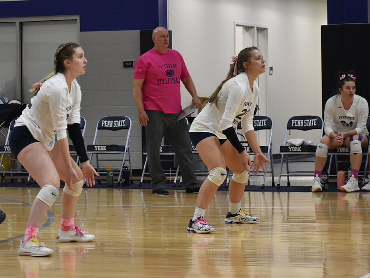 Two women's volleyball players on the court and one on the bench, male coach in the background wearing pink shirt.