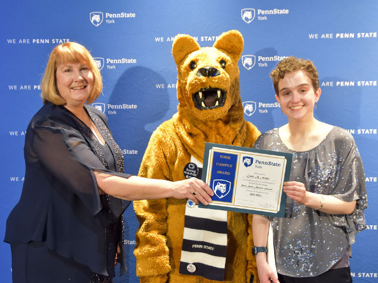 Famale presents an award to another a student, also female. Person in Nittany Lion costume between the two.