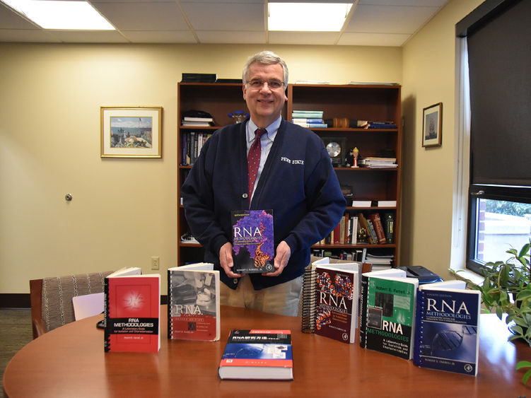 White older male wearing glasses with six books displayed on his desk.