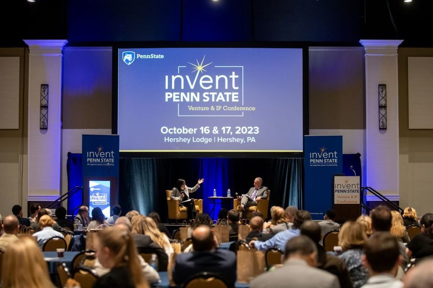 Dr. Karen Kim and Andrew Read speak sitting on a stage in front of an Invent Penn State Venture & IP Conference branded screen with a full audience in the foreground