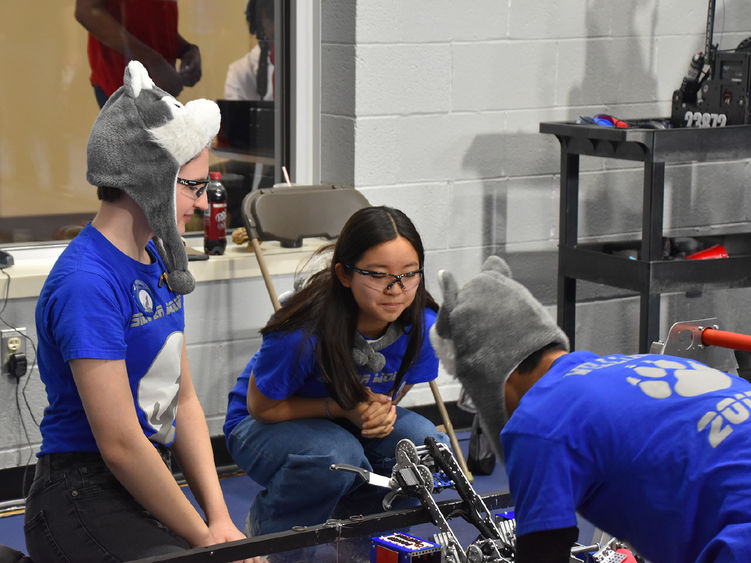 Three students work on a robot, one is wearing a wolf hat